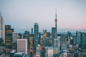 Photograph of a city skyline at dusk featuring numerous high-rise buildings with lights beginning to illuminate. Prominent CN Tower stands centrally, highlighting urban density and architectural variety in a metropolitan setting.
