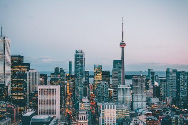 Photograph of a city skyline at dusk featuring numerous high-rise buildings with lights beginning to illuminate. Prominent CN Tower stands centrally, highlighting urban density and architectural variety in a metropolitan setting.