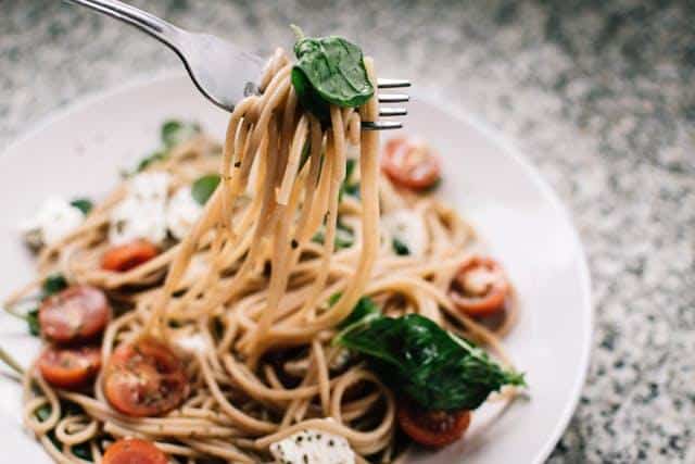 plate of spaghetti pasta mixed with cherry tomatoes, spinach leaves, and small pieces of cheese on a speckled surface