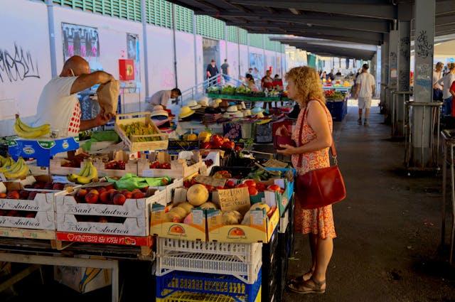 outdoor market stall under a covered area, featuring a vendor arranging fresh fruits and vegetables while a customer in a patterned dress with a red purse browses
