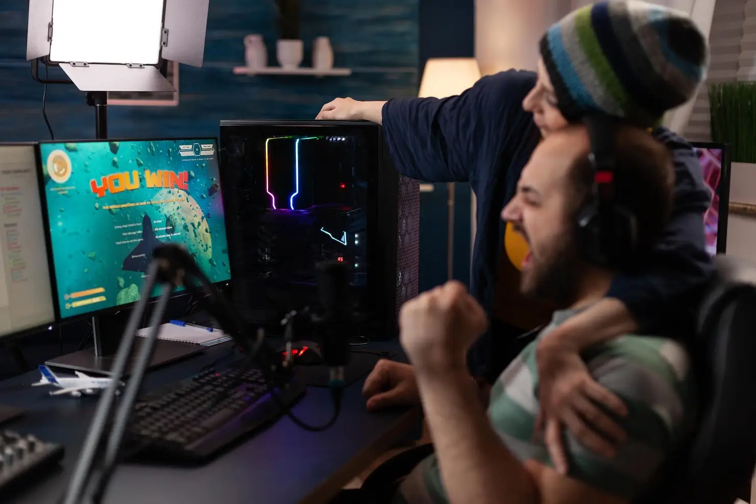 Two people celebrating gaming victory at a computer desk with colorful LED-lit PC tower