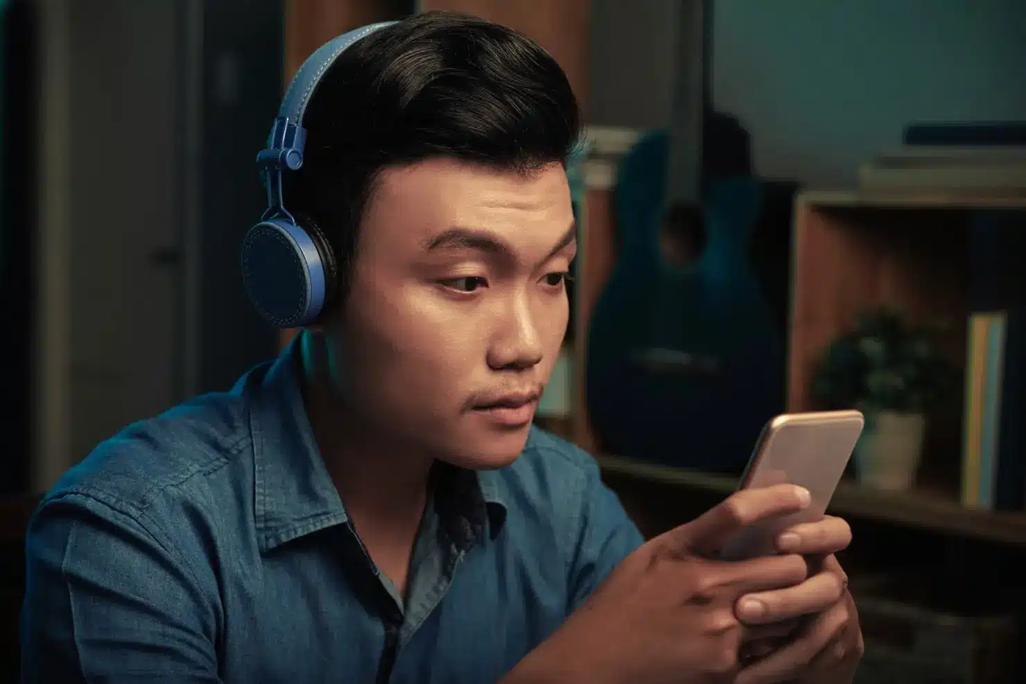 Man wearing blue headphones looking at smartphone screen in cozy room with guitar in background