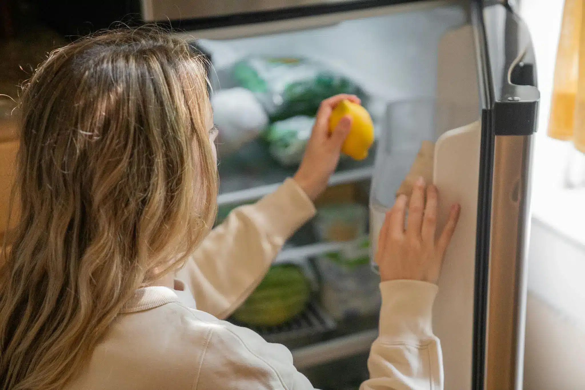 Person reaching for a lemon inside an open refrigerator in a brightly lit kitchen
