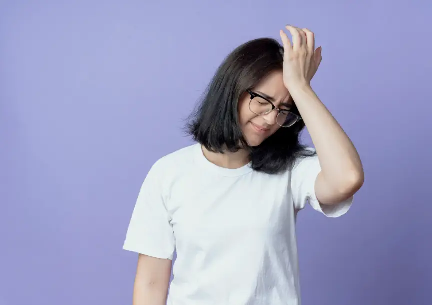 Woman with glasses in white t-shirt holding head in frustration against purple background