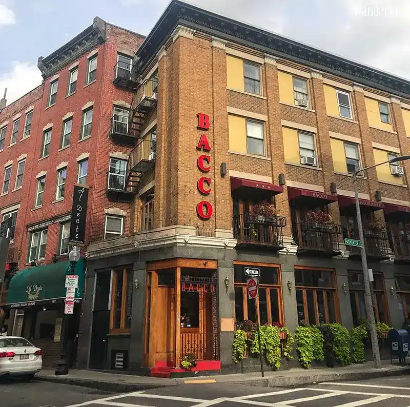 Three-story brick building with red "BACCO" sign on corner, featuring outdoor balconies and plants