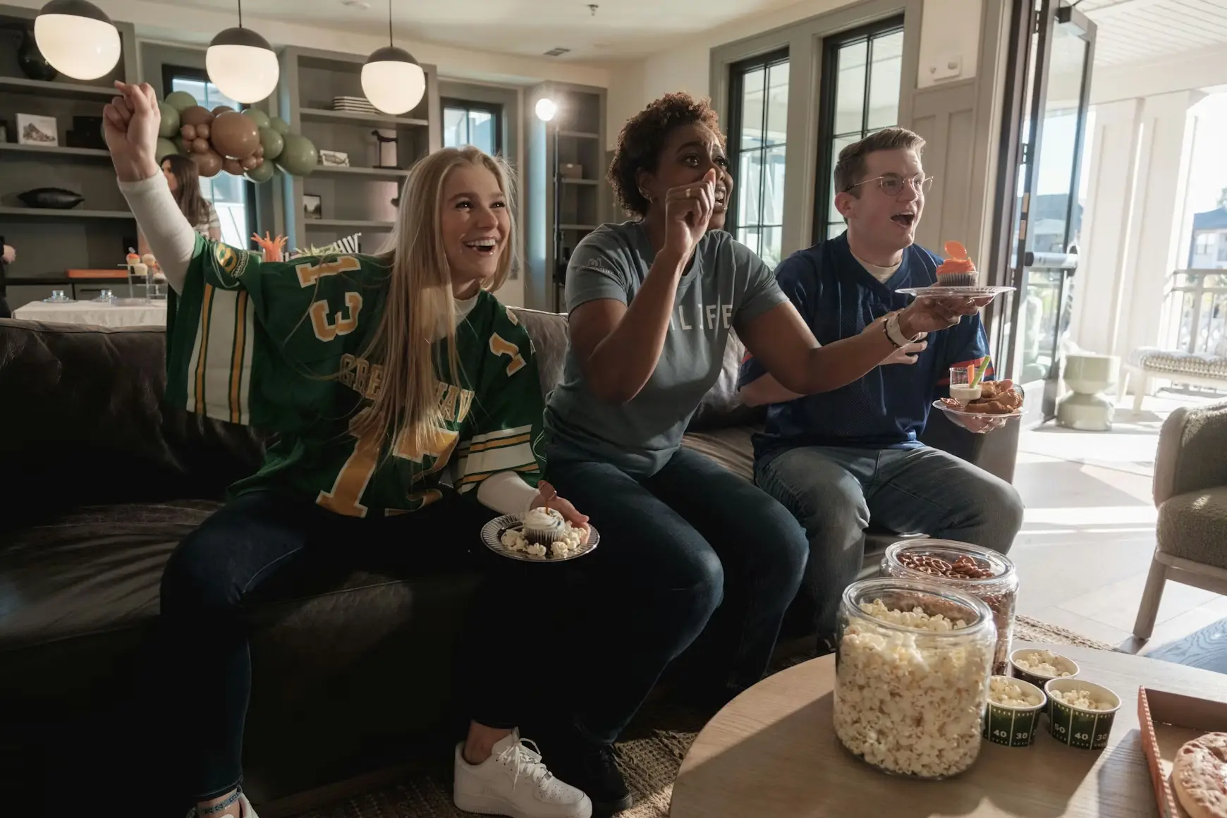Three people cheering while watching sports on TV in a living room with snacks