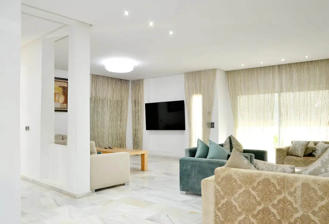 Modern living room with neutral sofas, wall-mounted TV, and sheer curtains under bright lighting