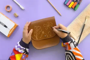 Person drawing floral design on brown leather purse with markers on purple surface