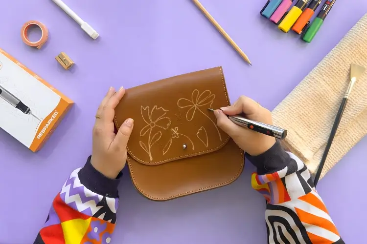 Person drawing floral design on brown leather purse with markers on purple surface