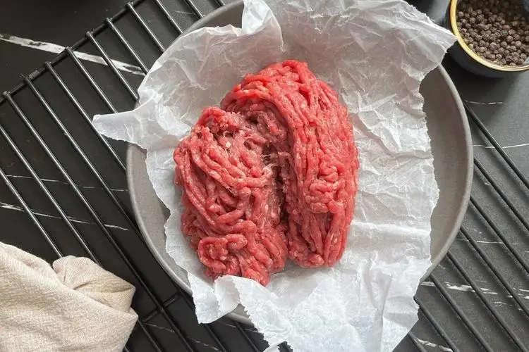 Raw ground beef on parchment paper in gray ceramic bowl on dark countertop