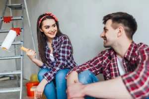 Couple in plaid shirts sitting by ladder with paint roller in hand