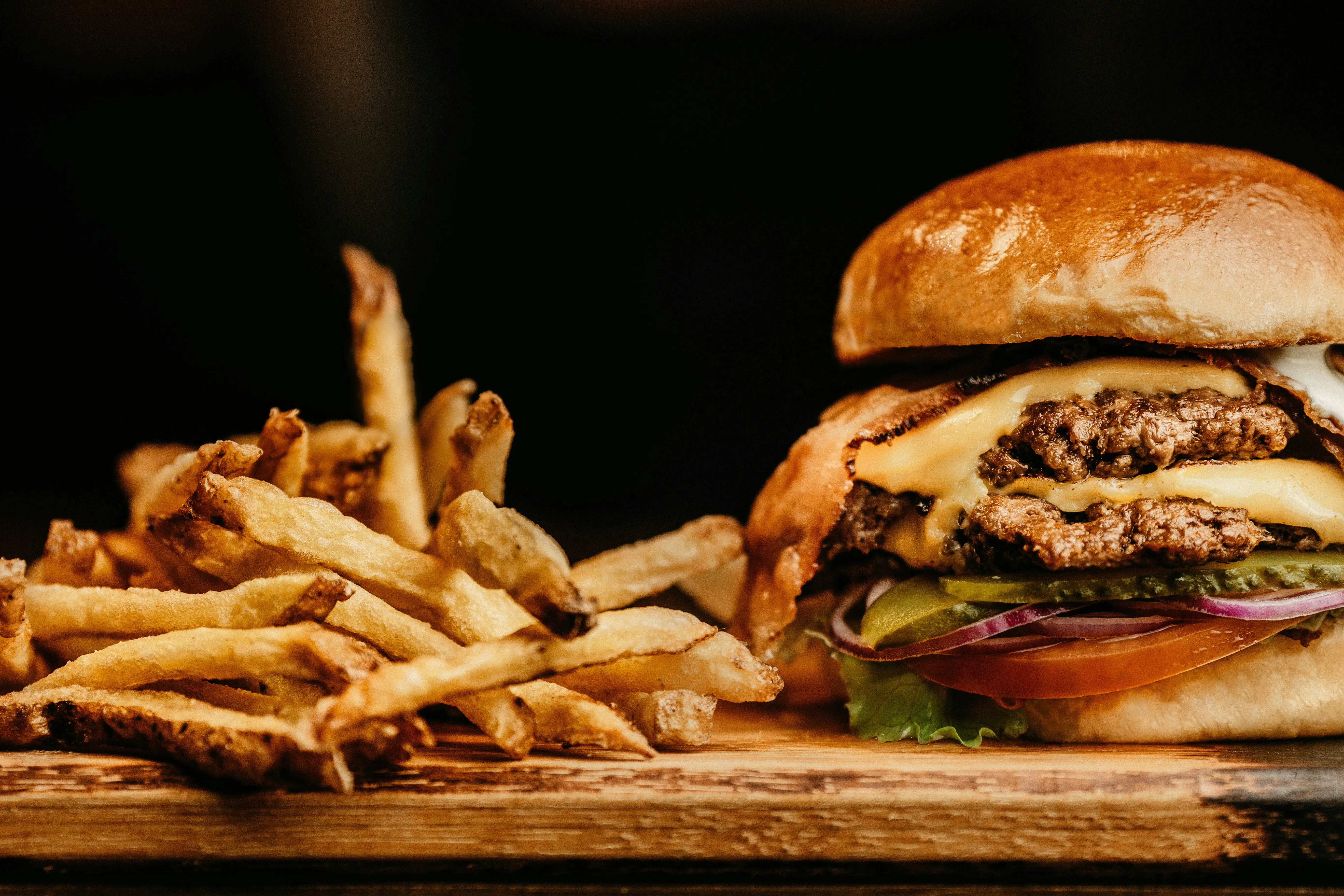 Juicy cheeseburger with bacon and fresh fries on wooden board in dimly lit setting