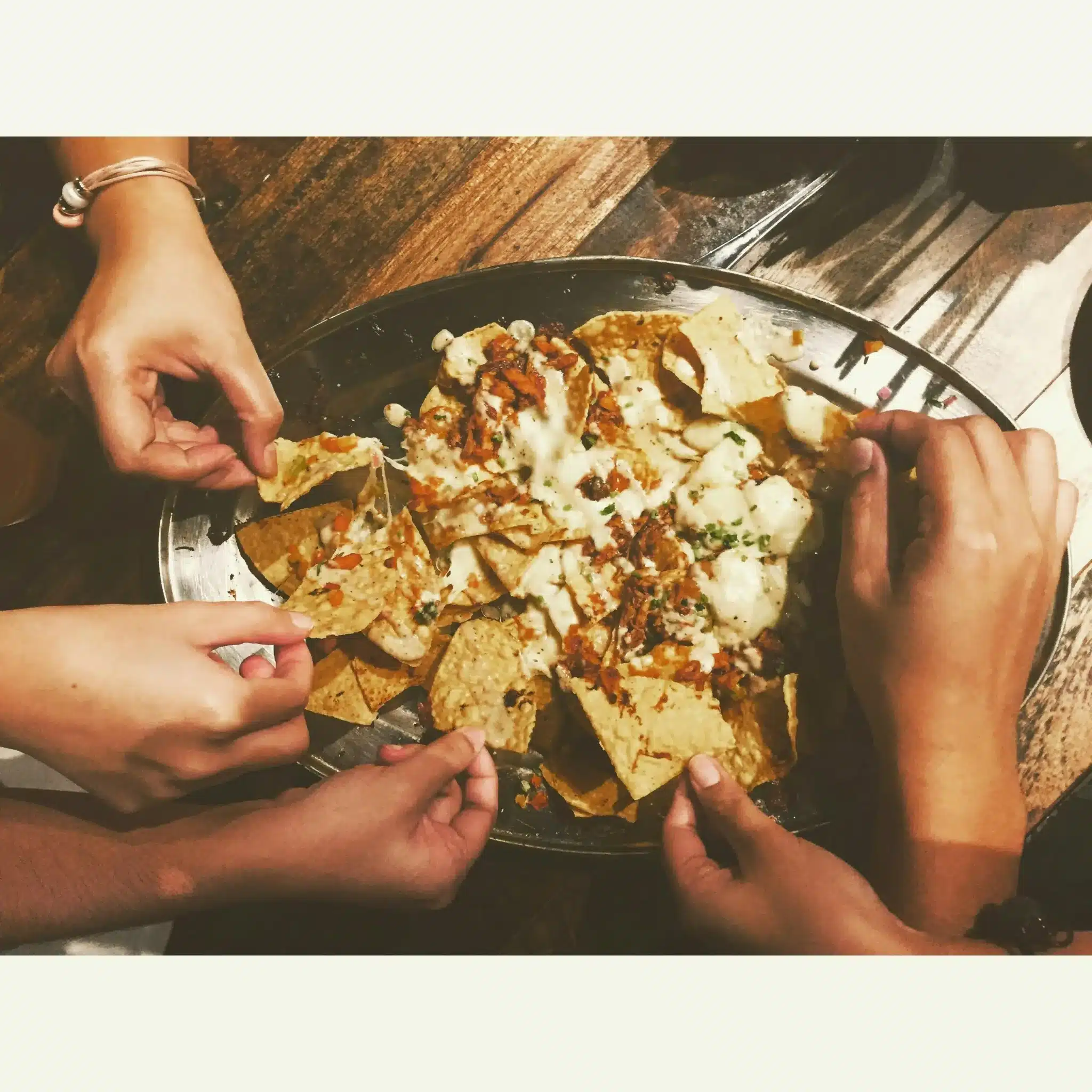 Hands reaching for nachos with melted cheese on metal tray on wooden table