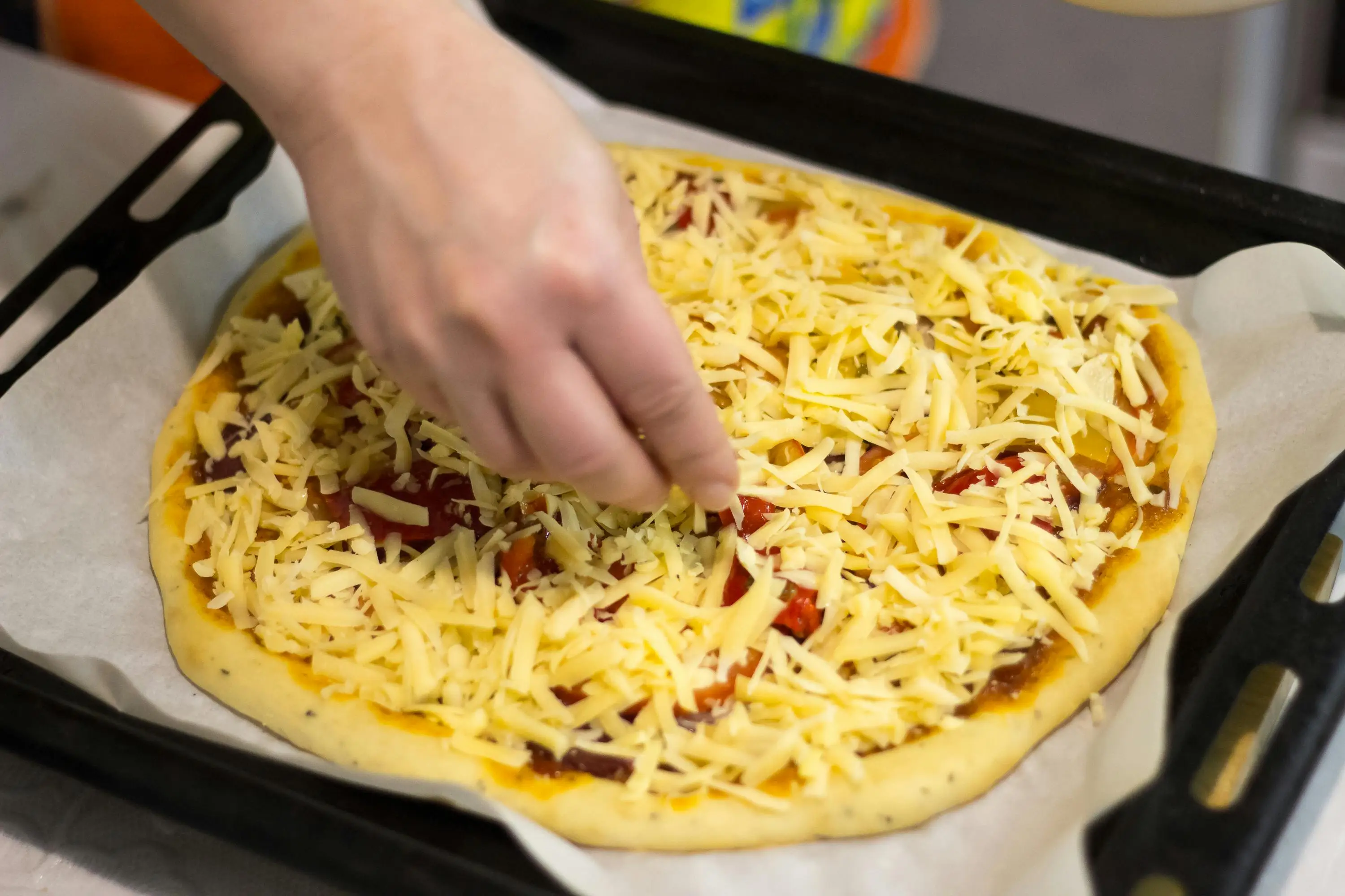 Hand adding grated cheese to unbaked pizza on parchment-lined baking tray