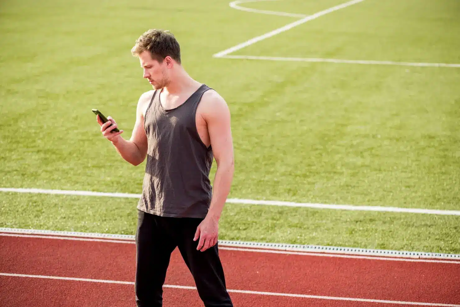 Man in tank top looking at smartphone on outdoor track under bright sunlight