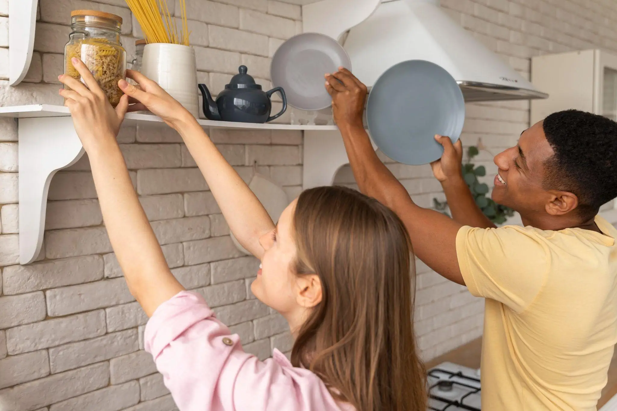 Couple organizing kitchen shelves with blue plates and jar of pasta on white brick wall