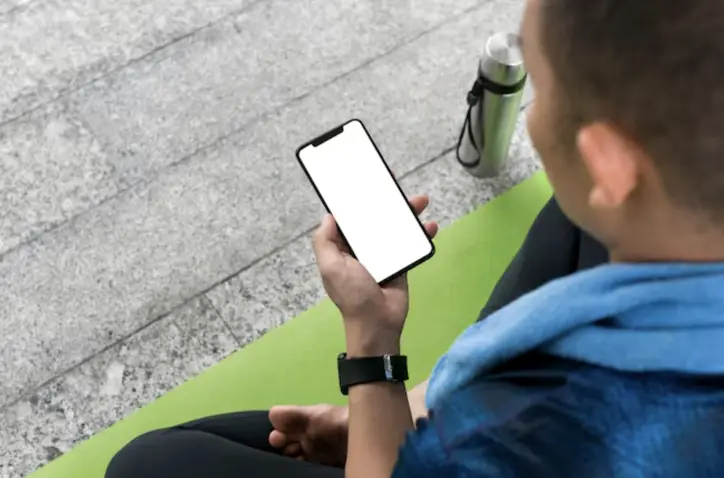 Person sitting on green yoga mat holding smartphone with blank screen outdoors