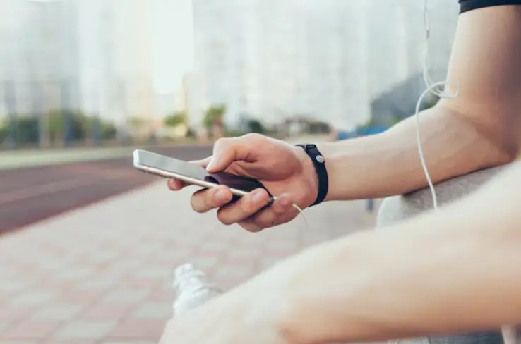 Person holding smartphone with water bottle and earphones on sunny outdoor track