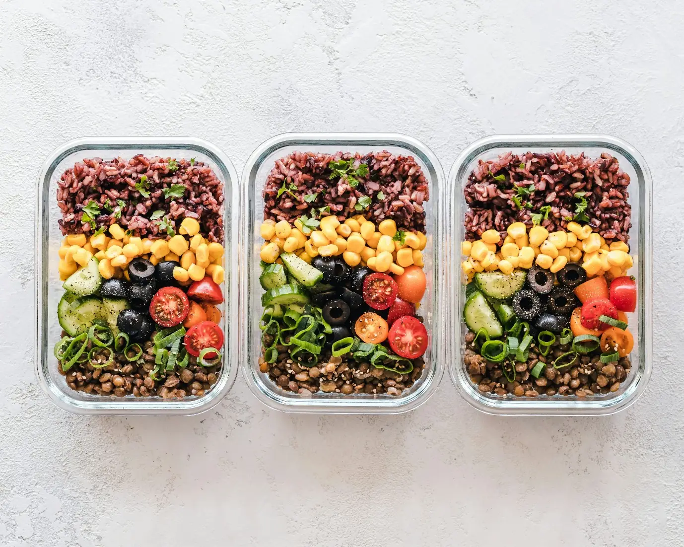 Glass meal prep containers with quinoa, corn, olives, cucumbers, and tomatoes on light textured surface
