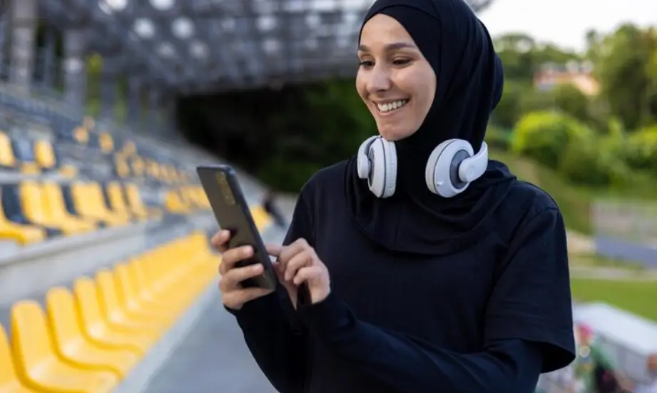 Young woman in hijab using smartphone with headphones in outdoor sports stadium