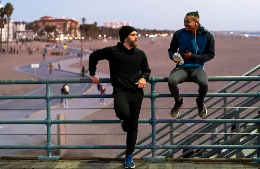 Two men talking on a beach boardwalk railing at sunset with blurred cityscape background