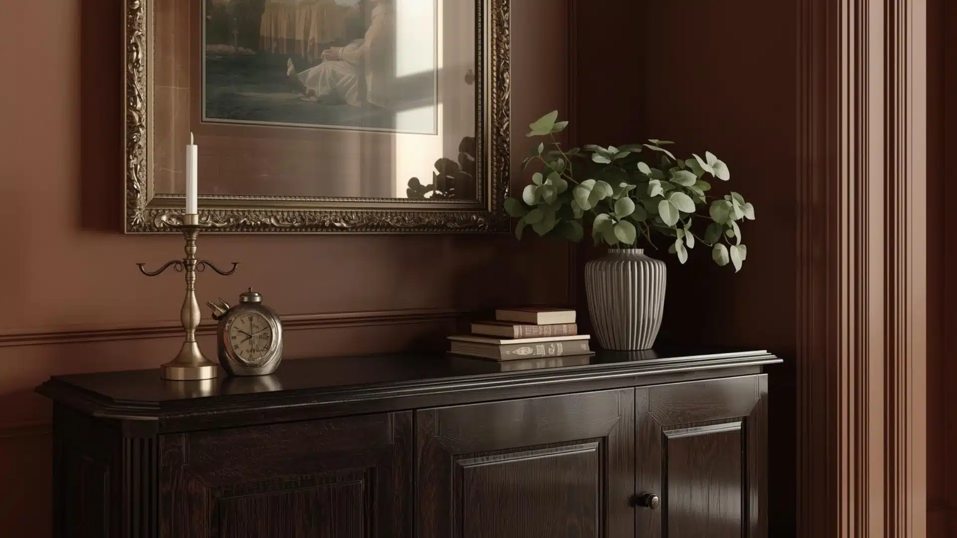 Modern Victorian sideboard with ornate mirror, brass candleholder, stacked books, and potted plant in warm light.