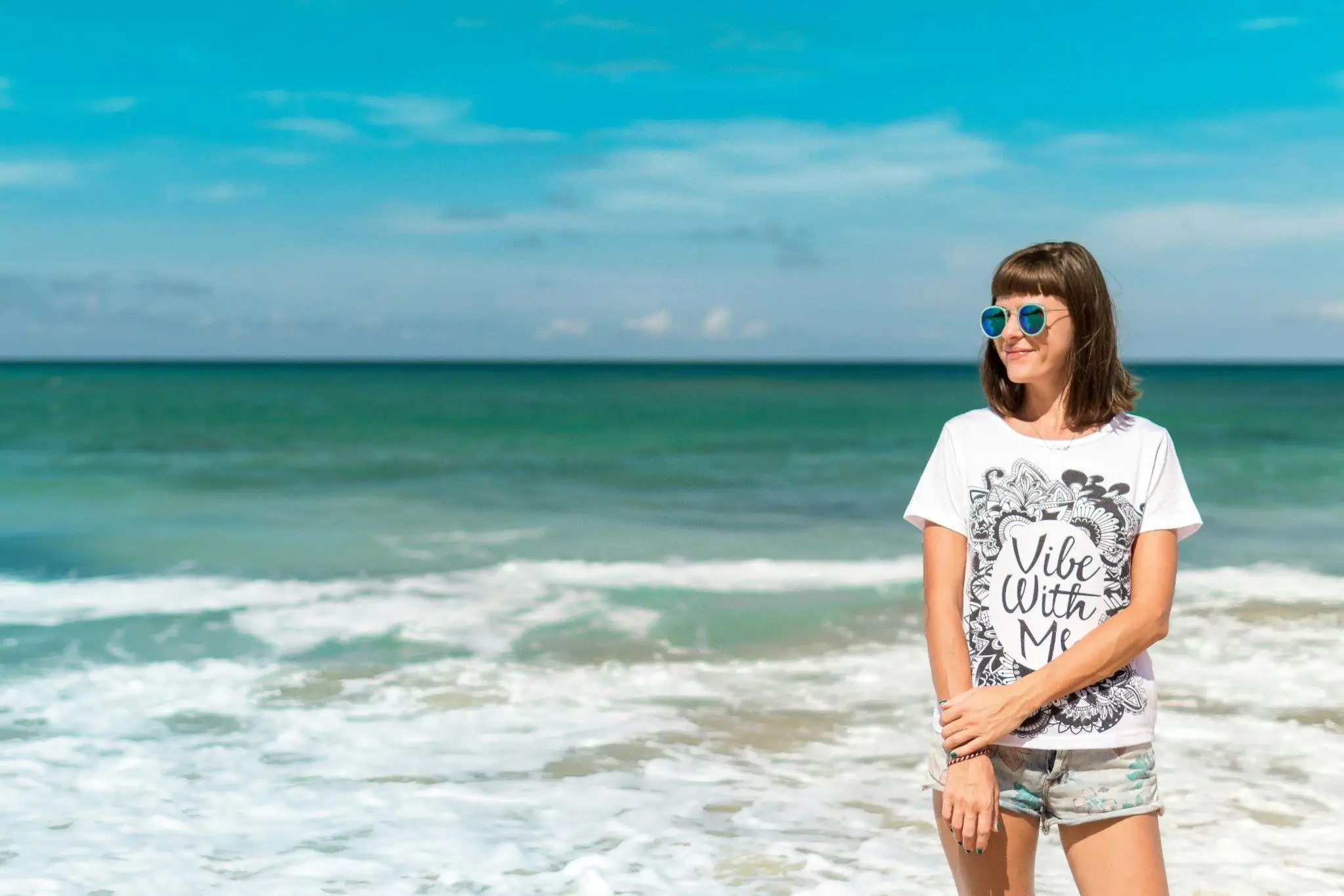 Woman in patterned white t-shirt and sunglasses standing on sunny beach with ocean in background