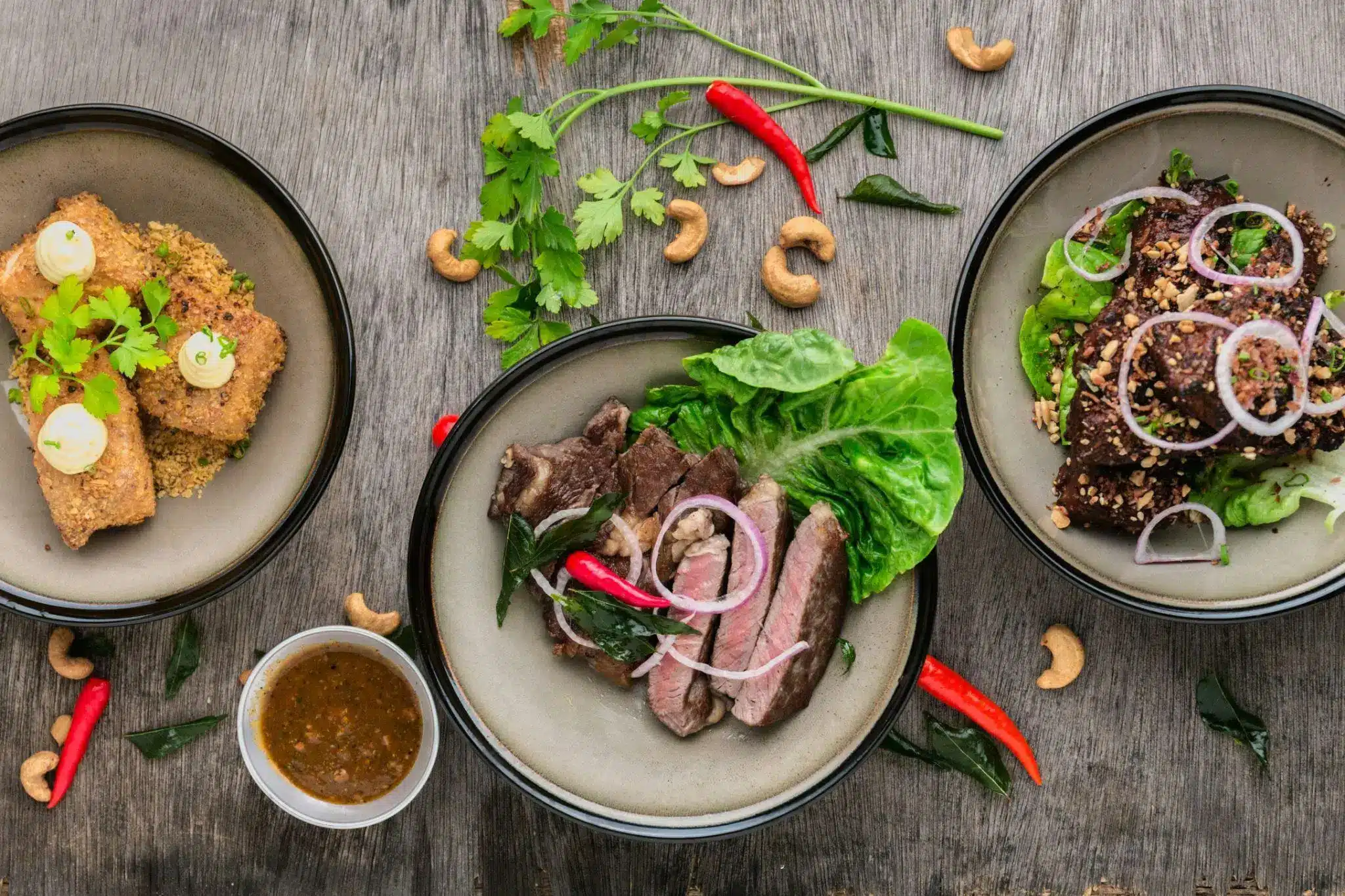 Assorted dishes with grilled meat, lettuce, nuts, and red chili on a wooden table