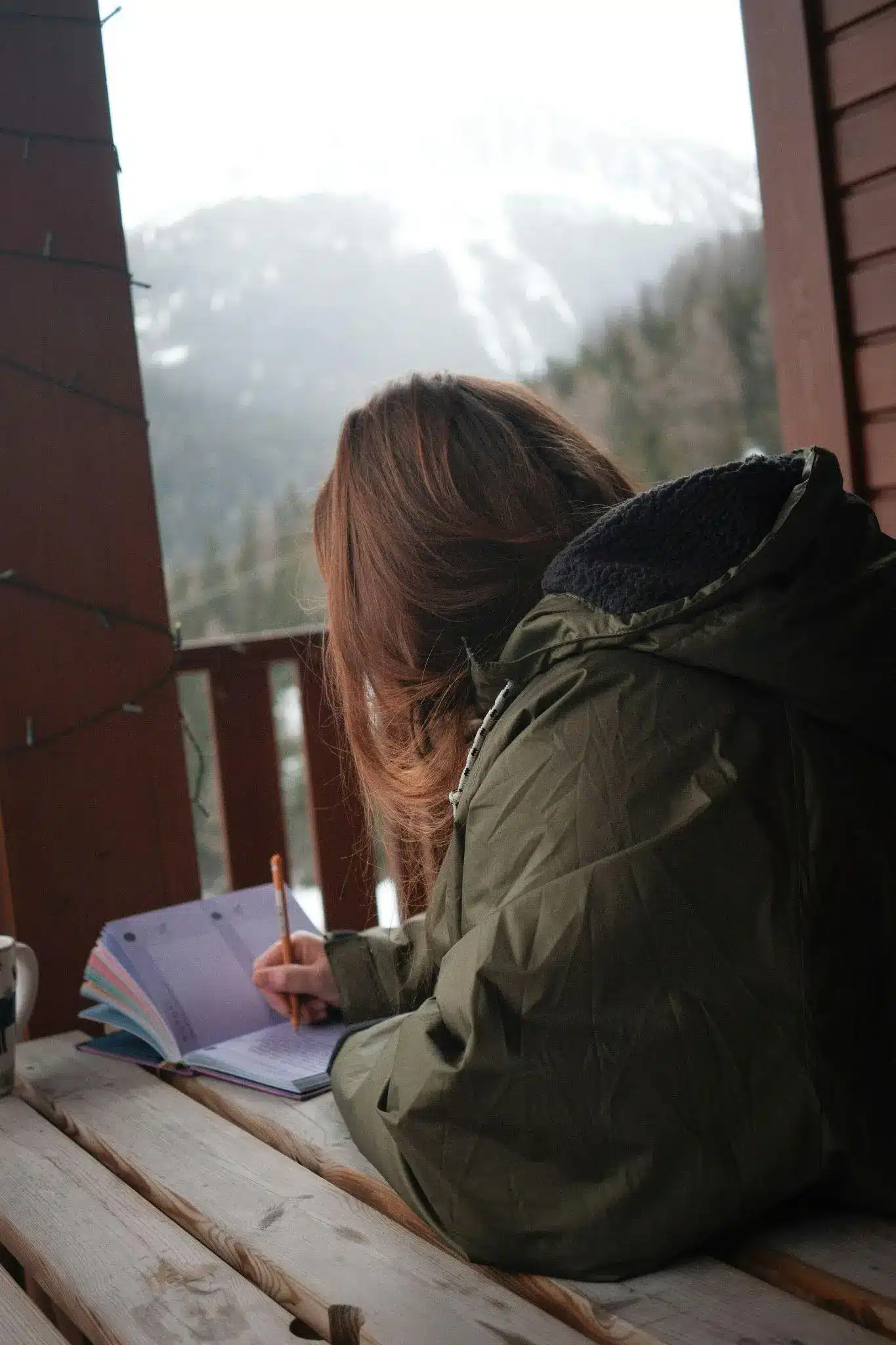 Person writing in a journal on a wooden balcony overlooking snowy mountains