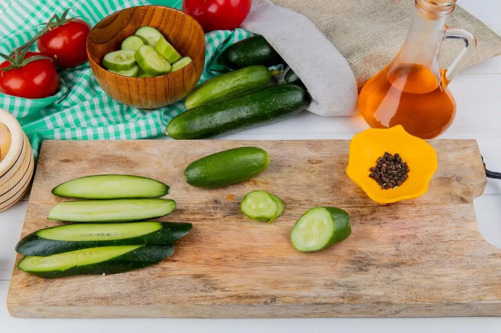 Sliced cucumbers on wooden cutting board with tomatoes, oil, and peppercorns nearby