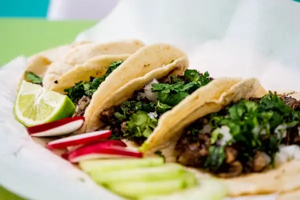Tacos filled with grilled meat and cilantro on a white plate with lime wedges and radish slices