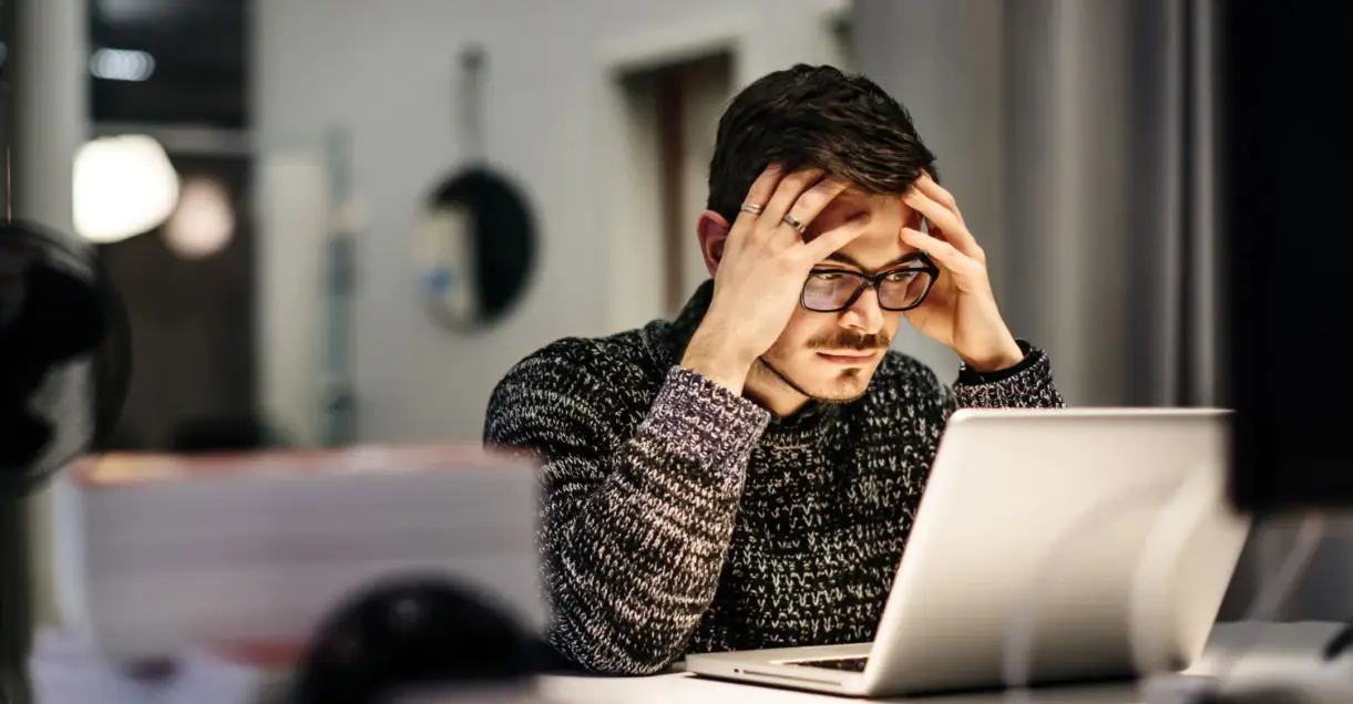 Man with glasses looking stressed while using laptop in dimly lit office