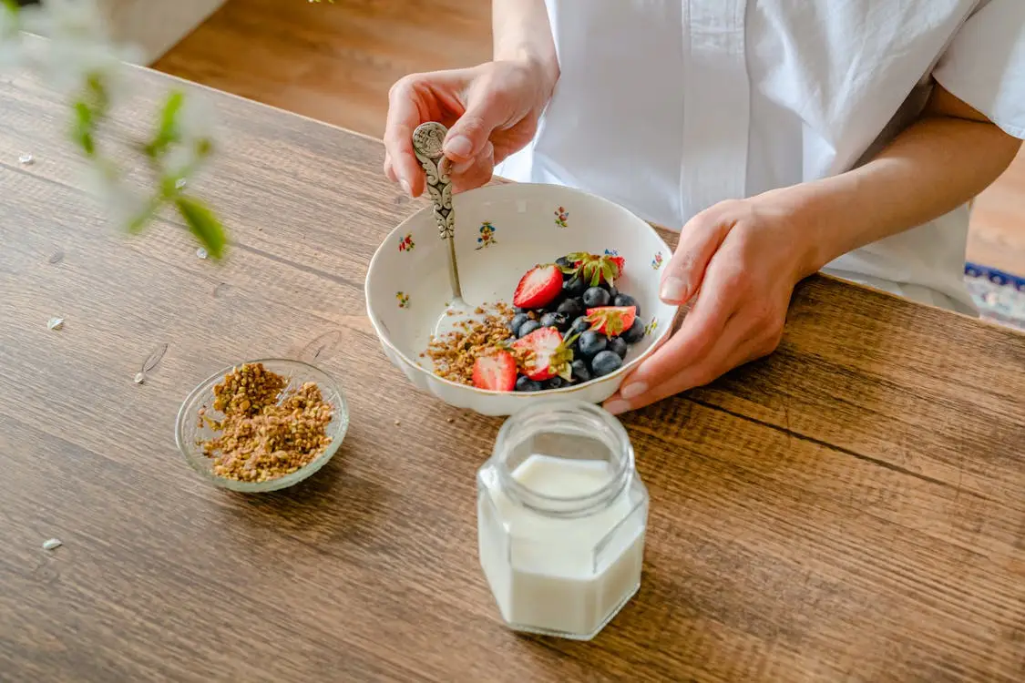 Person holding bowl of berries and granola on wooden table with milk and small dish nearby