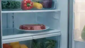 Raw steak on white plate inside a refrigerator with fresh vegetables surrounding it