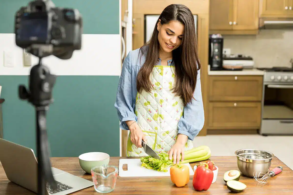 Woman chopping celery on a wooden counter in a bright kitchen with camera setup