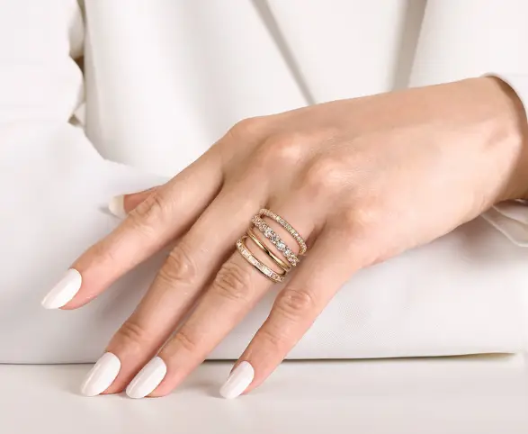 Hand with white manicure wearing layered gold rings and diamonds on a white fabric background