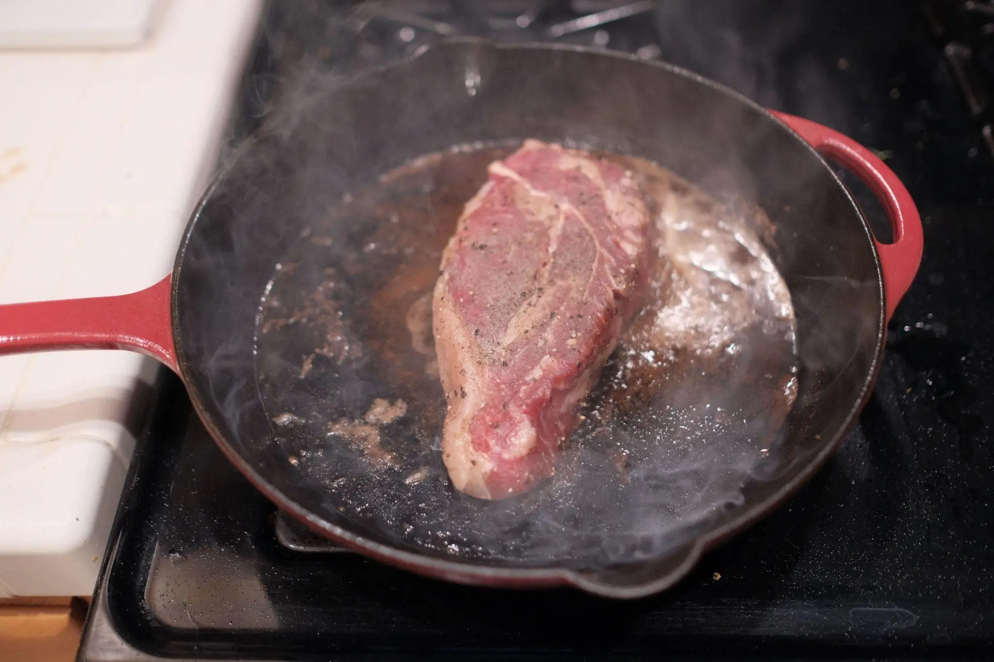 Steak searing in a red cast iron skillet with steam rising on stovetop