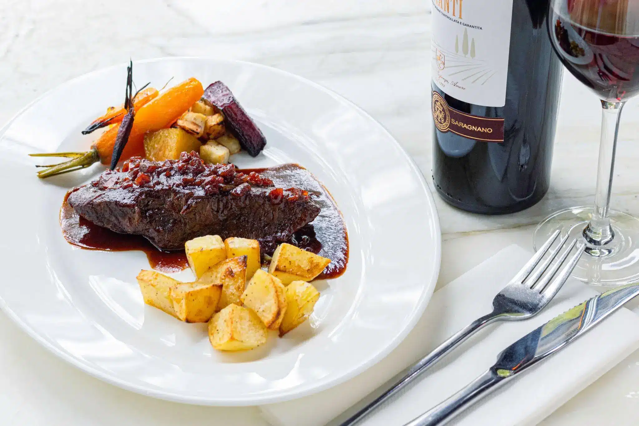 Gourmet beef dish with roasted vegetables and potato wedges on white plate near red wine bottle