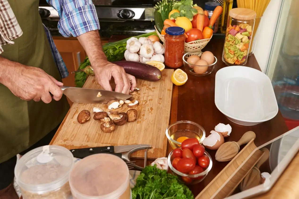 Person slicing mushrooms on wooden cutting board in kitchen with fresh vegetables and eggs nearby
