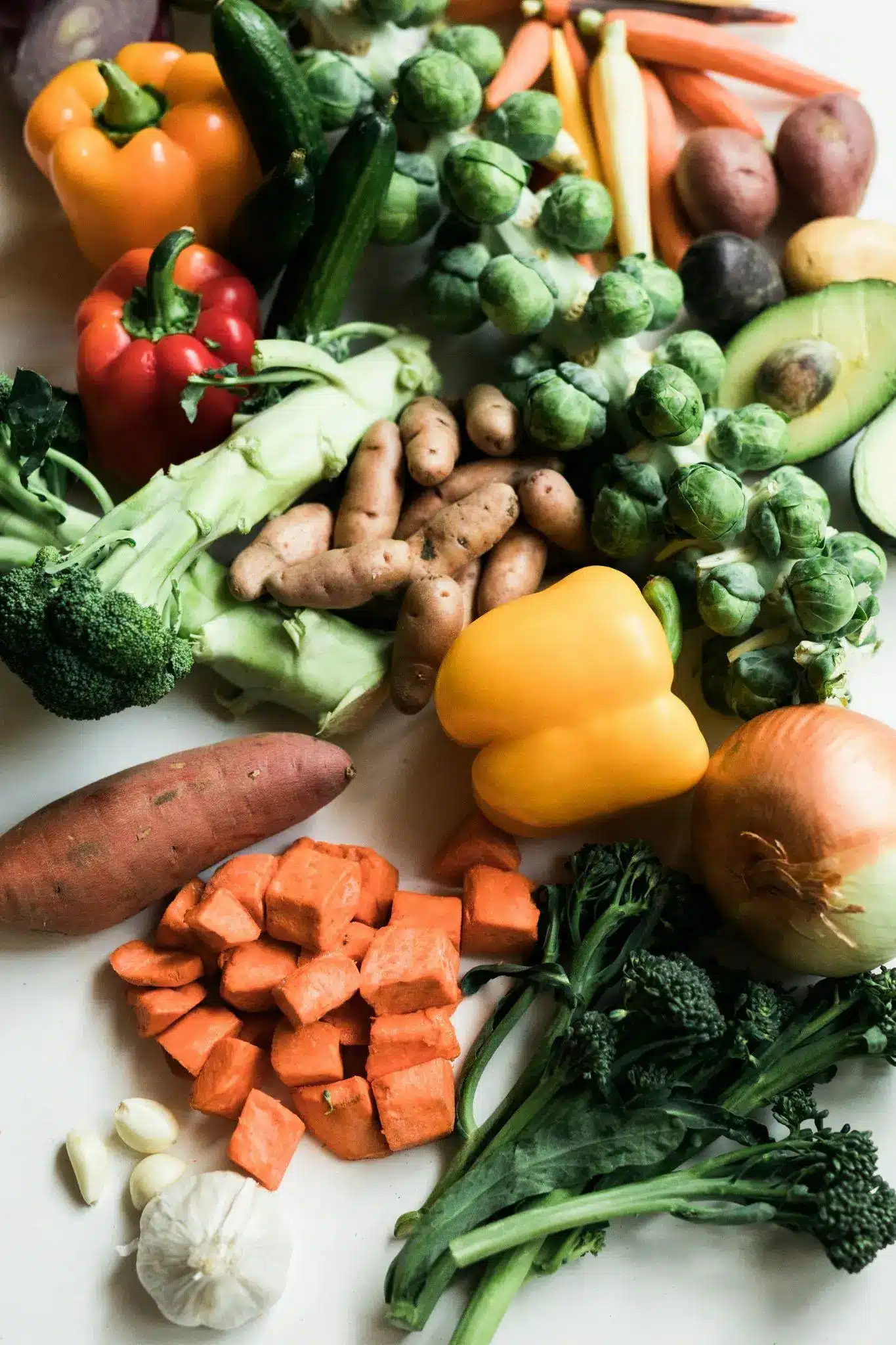 Assorted fresh vegetables including peppers, broccoli, and sweet potatoes on a white surface