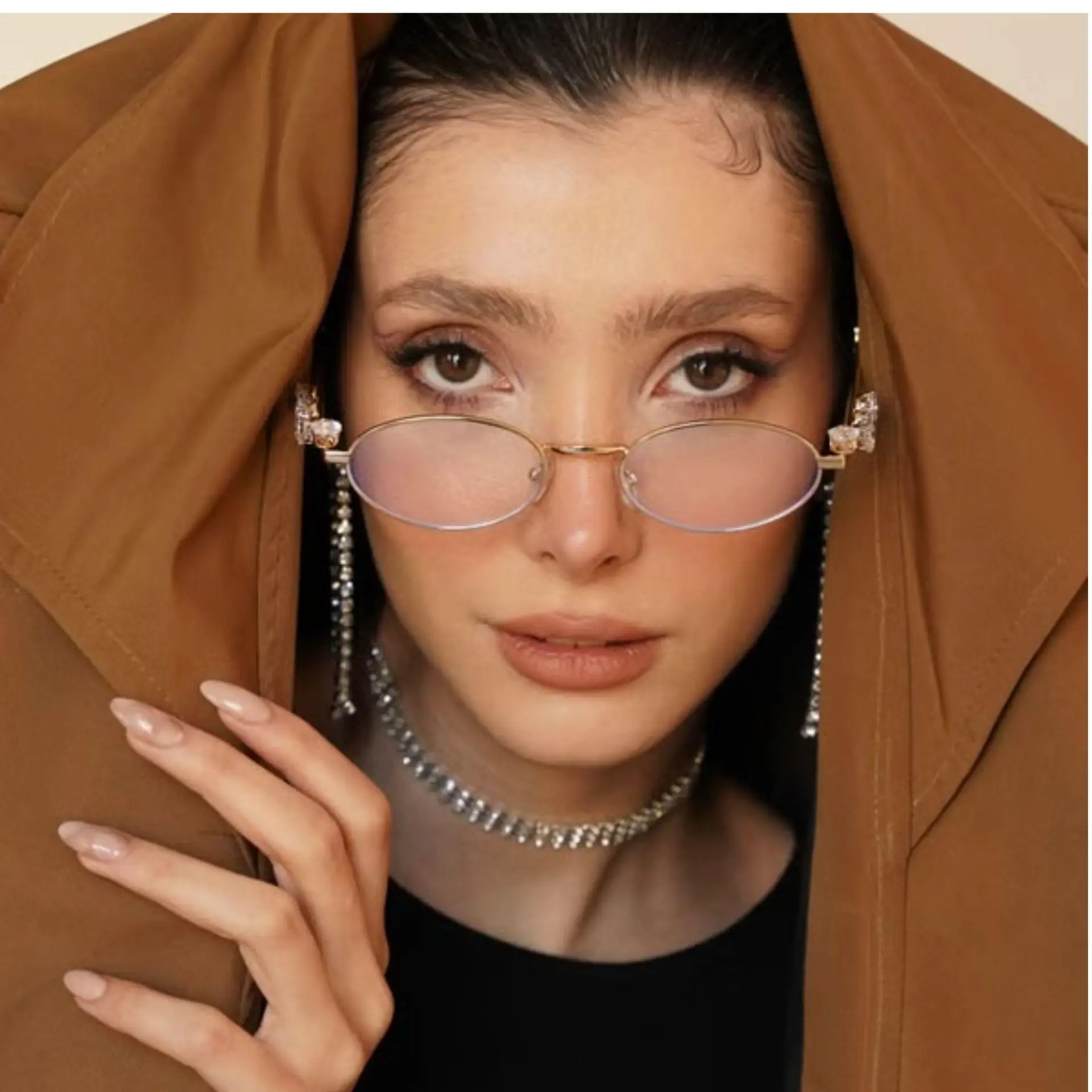 Person wearing stylish glasses and crystal jewelry under brown coat indoors