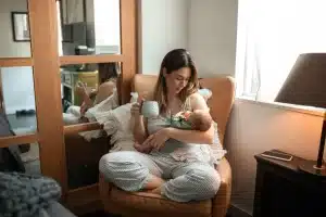 Woman in striped pajamas holding baby and mug in cozy living room chair