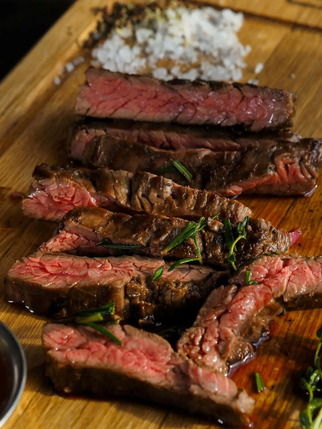Sliced medium-rare steak with herbs and coarse salt on a wooden board