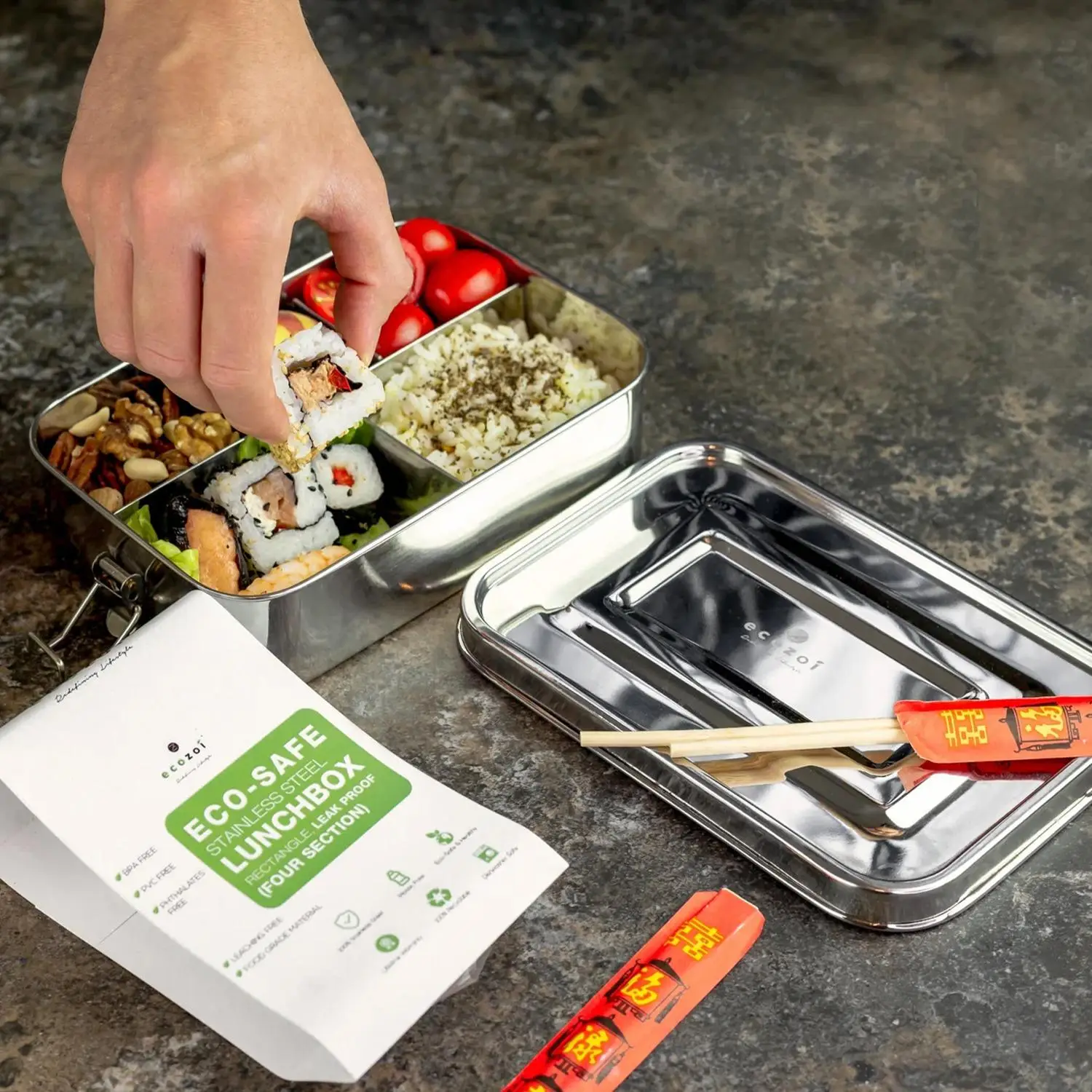 Hand placing sushi in a stainless steel lunchbox with compartments on a stone countertop