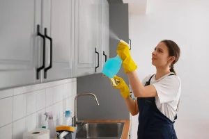 Person wearing gloves cleaning kitchen cabinets with spray bottle above sink