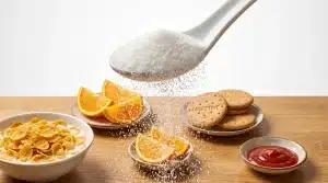 Large spoon sprinkling sugar over cornflakes, orange slices, cookies, and ketchup on wooden table