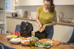 Person preparing vegetables in air fryer on wooden kitchen counter with fresh produce and olive oil