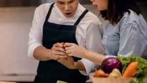 Chef in dark apron and woman in denim shirt examining injury in bright kitchen setting