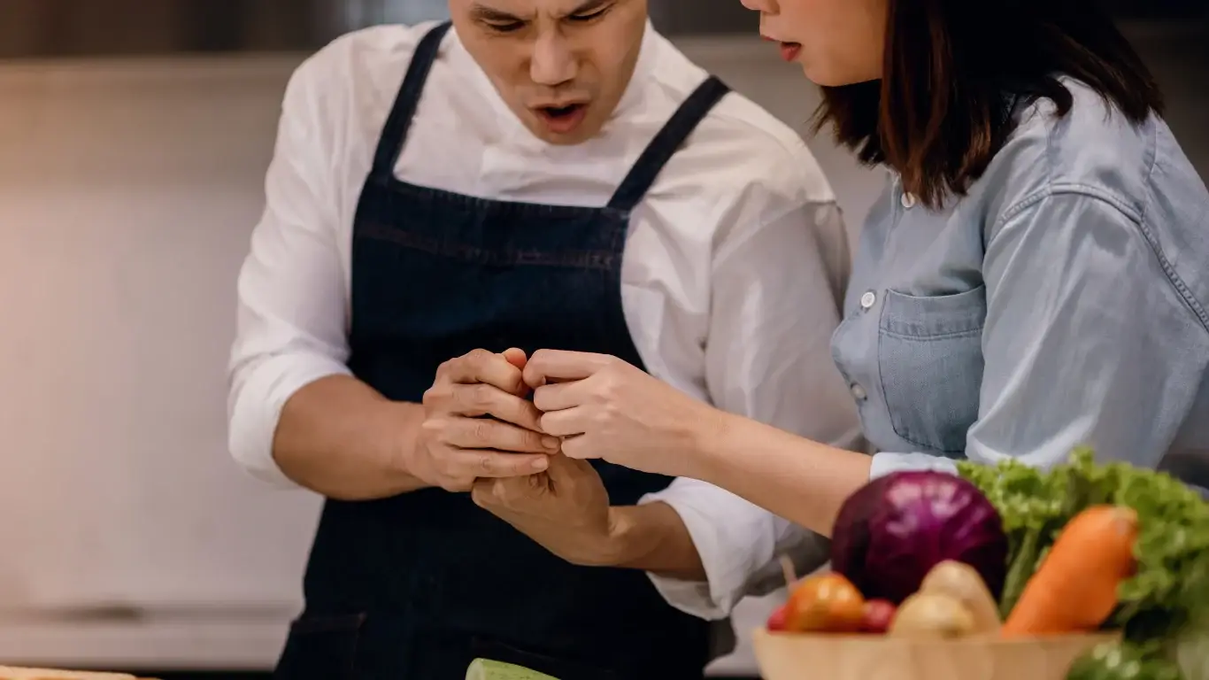Chef in dark apron and woman in denim shirt examining injury in bright kitchen setting