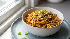 Noodles topped with sesame seeds and green onions in a ceramic bowl by a window